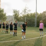 Treinadora observa jogadoras de futebol durante um treino ao ar livre em um campo ensolarado.