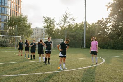 Treinadora observa jogadoras de futebol durante um treino ao ar livre em um campo ensolarado.