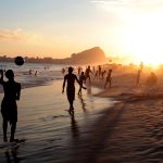 Pessoas jogam futebol na areia à beira-mar, em uma praia do Rio de Janeiro.