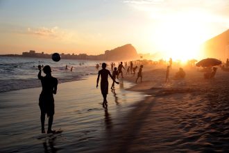 Pessoas jogam futebol na areia à beira-mar, em uma praia do Rio de Janeiro.